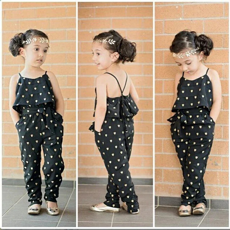 Little girl in black jumpsuit with gold heart print, floral headband, against brick wall