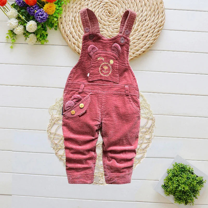 Pink corduroy baby overalls with bear design, displayed on white wooden background