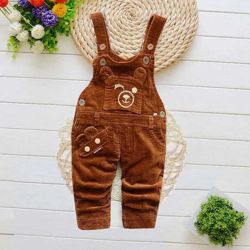 Brown corduroy baby overalls with bear face, buttons, on white wooden background with flowers.