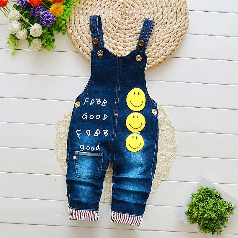 Blue denim baby overalls with smiley faces and 'good' print, styled on a white wooden background