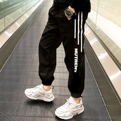 Black jogger pants with white stripes and NUTRENT logo, worn with white sneakers in a modern hallway