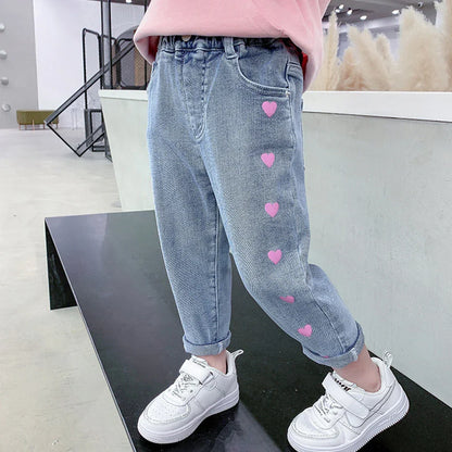 Kids' light blue jeans with pink heart designs and white sneakers in modern indoor setting