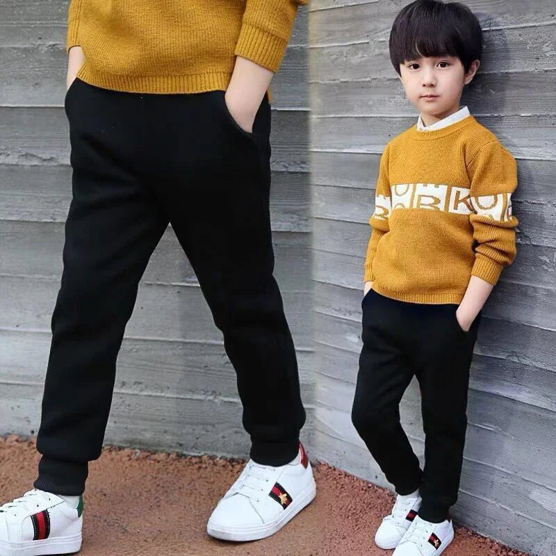 Boy in mustard sweater and black joggers with white sneakers posing by gray wall
