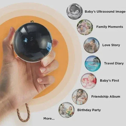 Hand holding a black photo locket sphere with icons for ultrasound, family, love, travel, baby, friends, and birthday memories.