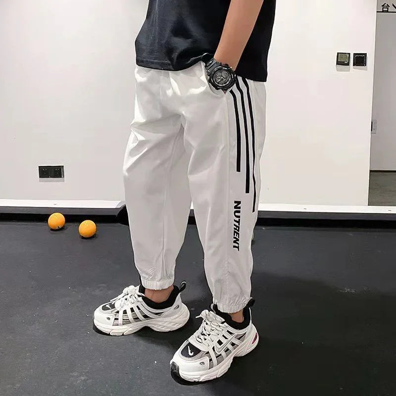 Man wearing white jogger pants with black stripes, chunky sneakers, and a wristwatch indoors