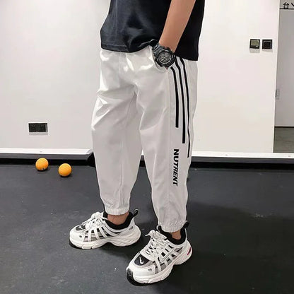 Man wearing white jogger pants with black stripes, chunky sneakers, and a wristwatch indoors